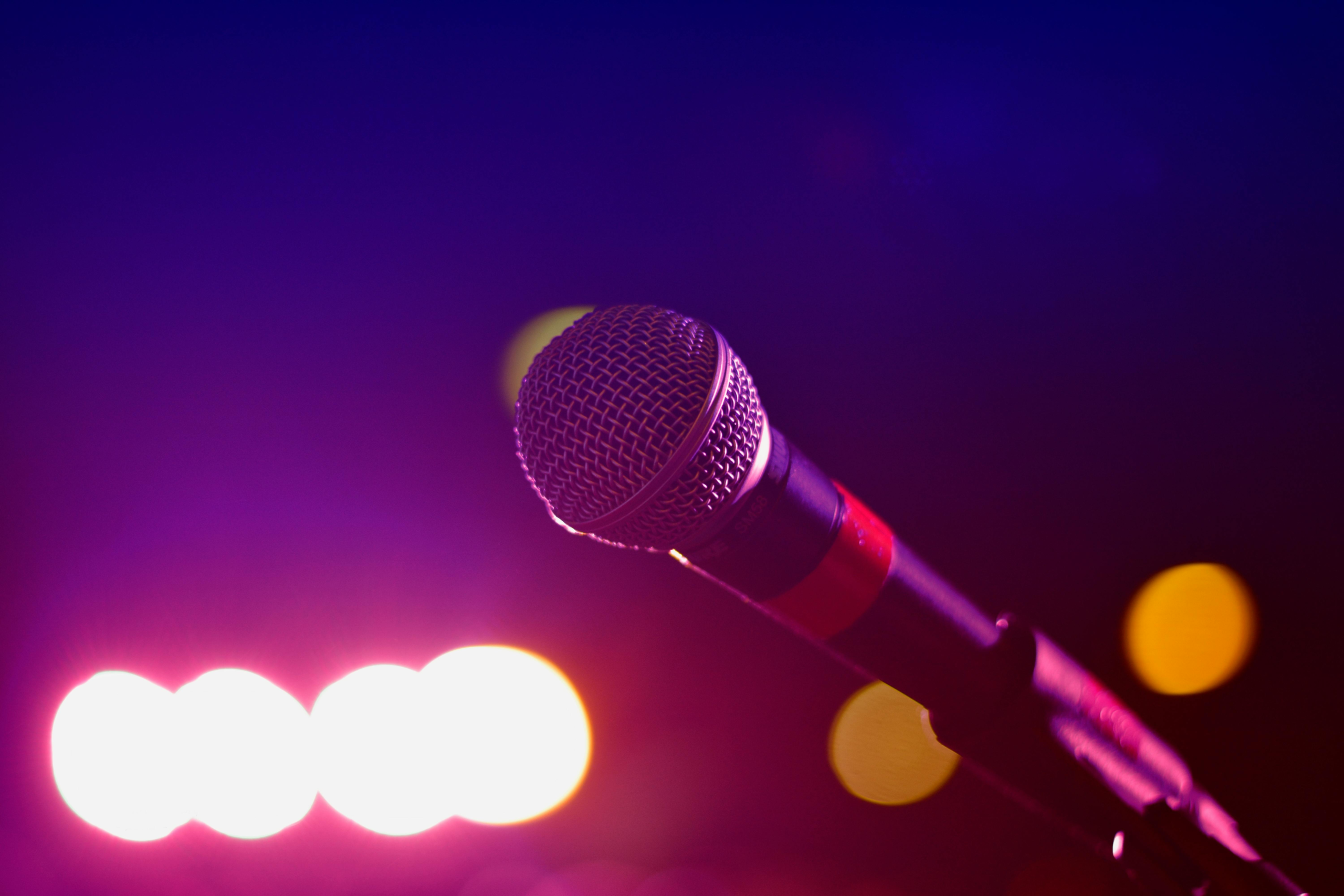 A handheld microphone photographed against a purple background, with blurred white stage lights and yellow bokeh effects in the background.