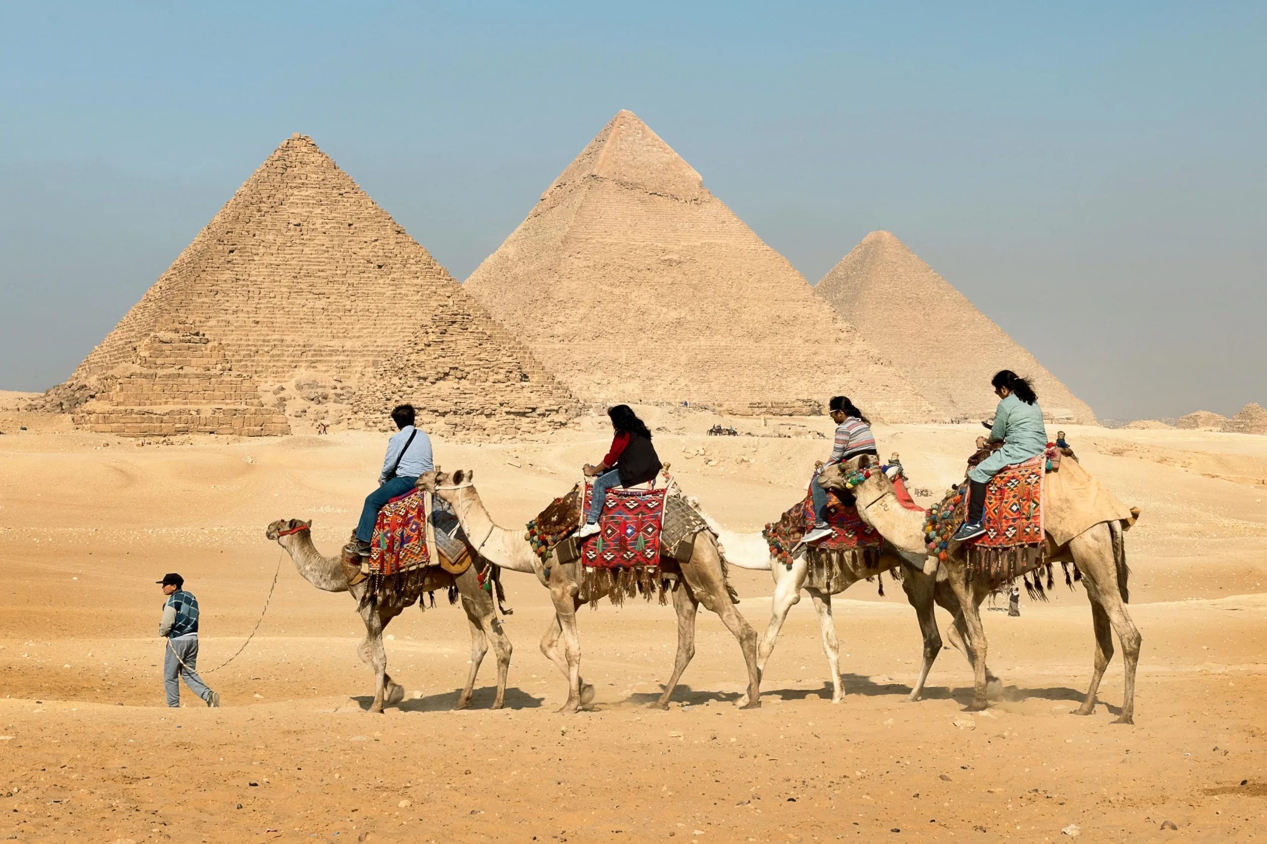 Tourists riding decorated camels in a line, led by a guide on foot, with the three Great Pyramids of Giza visible in the background against a clear blue sky in the Egyptian desert.