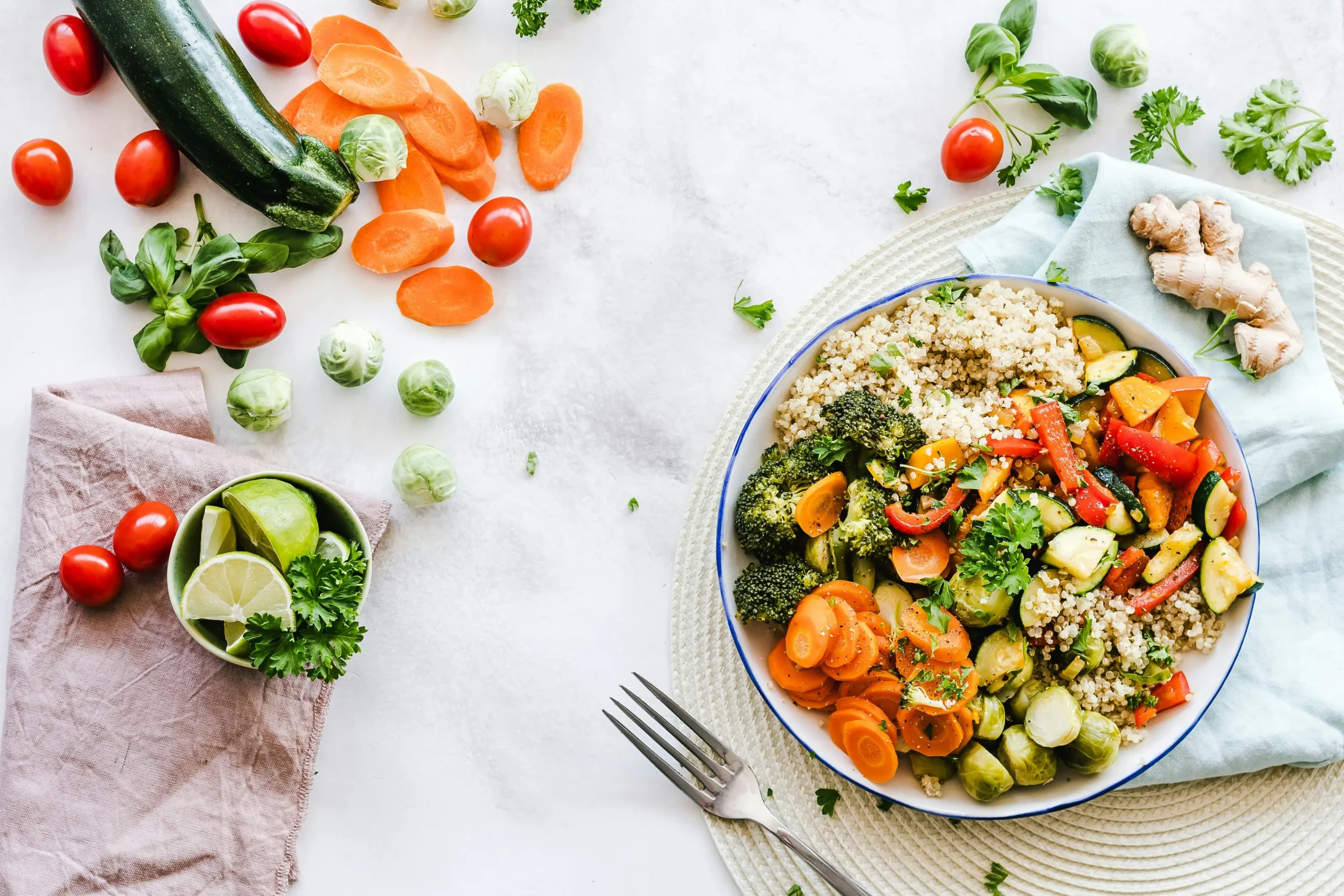 A healthy bowl with quinoa, roasted vegetables including broccoli, carrots, and Brussels sprouts, alongside fresh ingredients like cherry tomatoes, zucchini, lime, and ginger on a white marble surface.