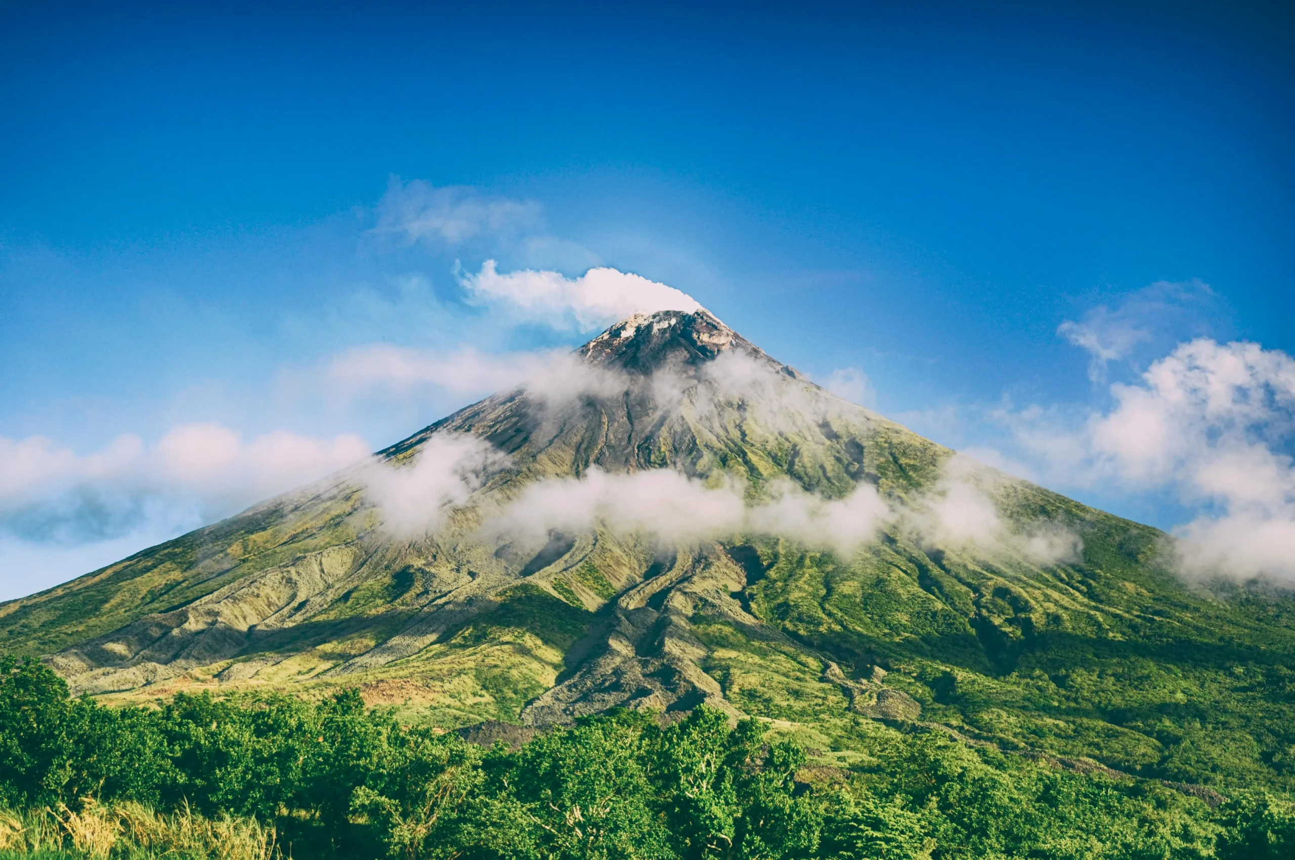 Mount Mayon volcano in the Philippines, a perfectly cone-shaped mountain with green vegetation on its slopes, white clouds wrapping around its peak, and a vibrant blue sky above.