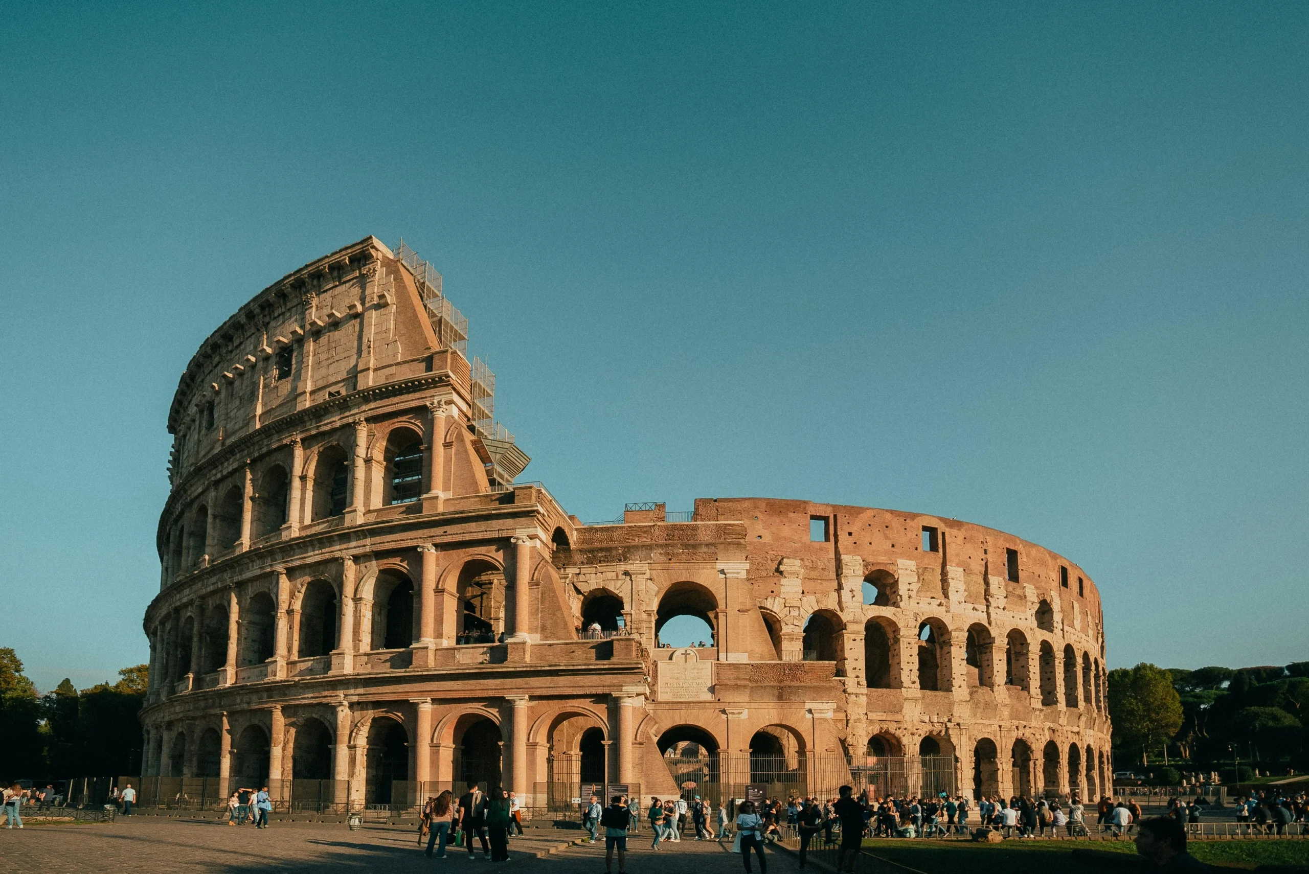 The Roman Colosseum in Rome, Italy, photographed during golden hour with tourists walking in front of the ancient amphitheater. The iconic stone arches and architecture are lit by warm sunlight against a clear blue sky.