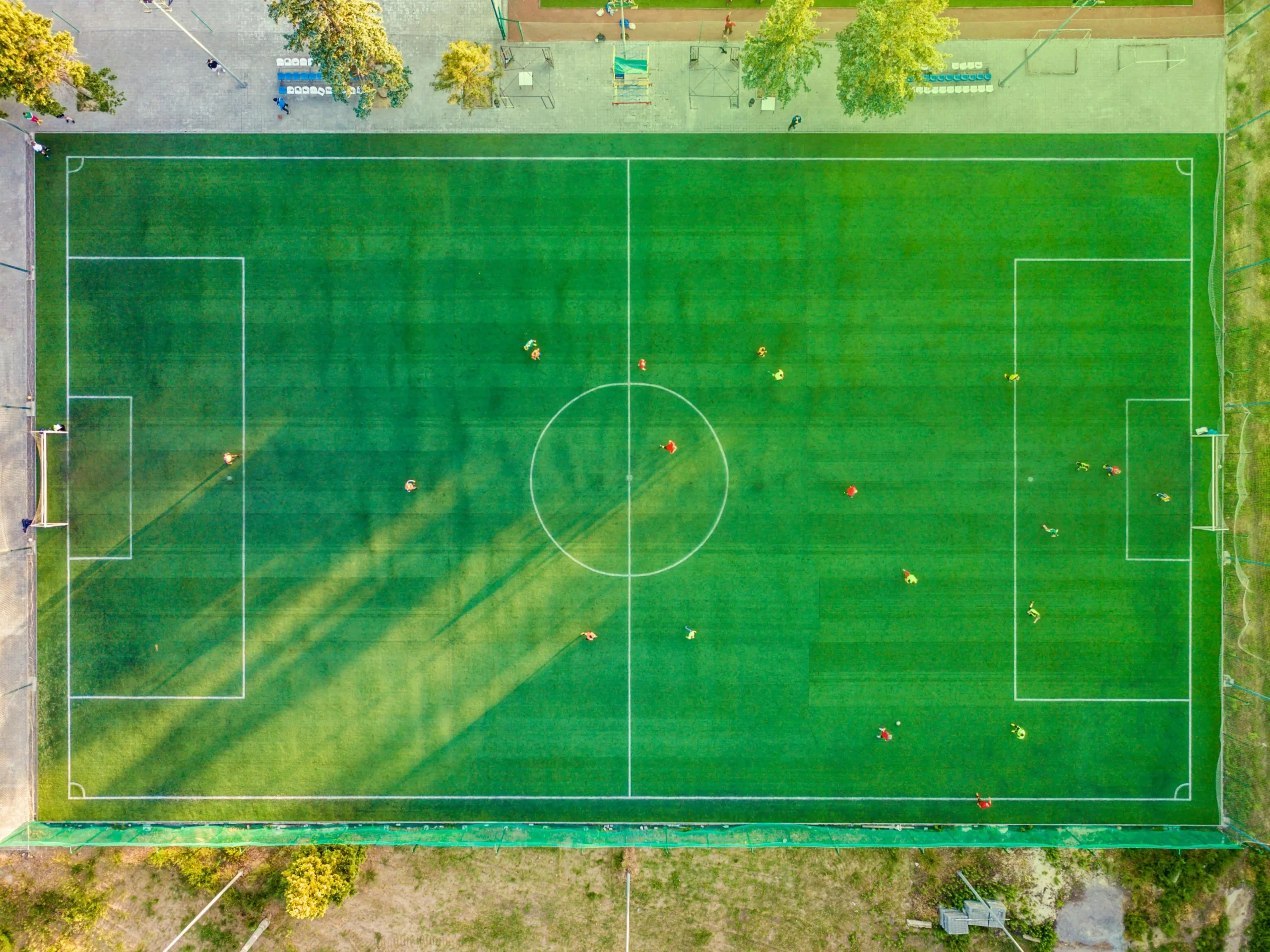 Aerial view of a green soccer field with white line markings, showing players scattered across the pitch during a match, surrounded by trees and facilities at the edges.